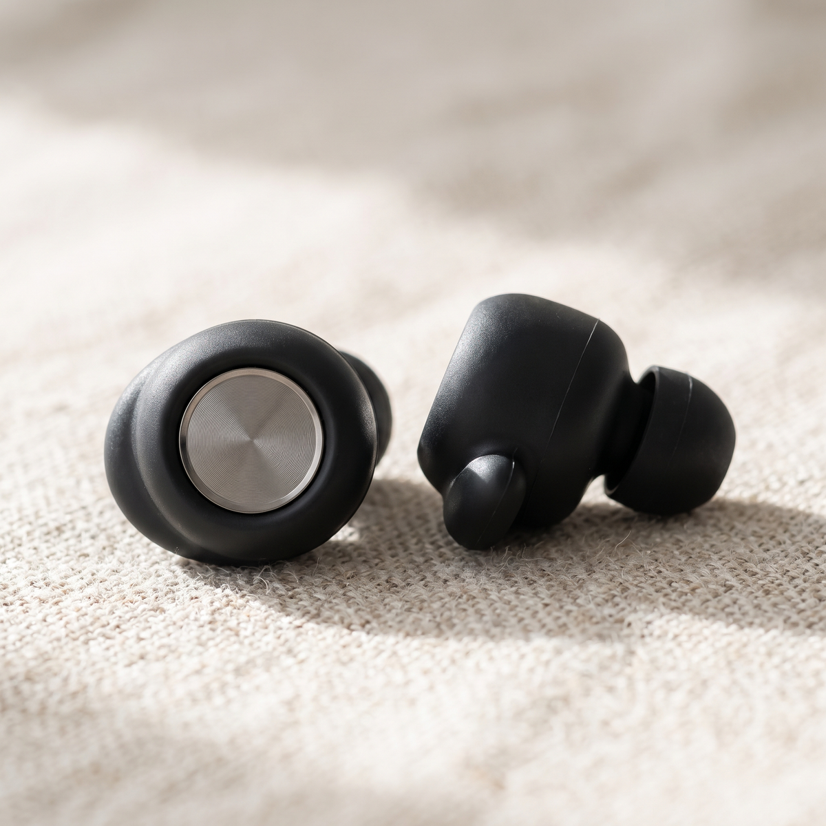 Close-up of two Vespré Grip earplugs on linen, brushed metal cap and soft silicone tip